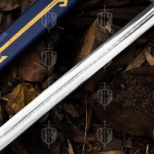 Pode incluir: Um close-up de uma l&acirc;mina de espada com desenhos gravados. A l&acirc;mina de a&ccedil;o polido est&aacute; em &acirc;ngulo, revelando detalhes da grava&ccedil;&atilde;o. Uma bainha azul com detalhes dourados &eacute; vis&iacute;vel no fundo, sugerindo um tema hist&oacute;rico ou de fantasia.