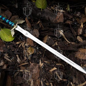 Pode incluir: Uma espada prateada com uma guarda detalhada com detalhes dourados e uma empunhadura envolta em azul e turquesa. A l&acirc;mina tem um emblema triangular perto da guarda. A espada &eacute; exibida sobre uma cama de folhas e galhos.