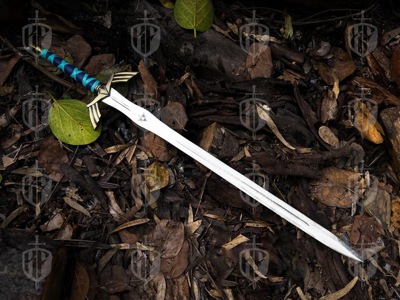 Pode incluir: Uma espada prateada com uma guarda detalhada com detalhes dourados e uma empunhadura envolta em azul e turquesa. A l&acirc;mina tem um emblema triangular perto da guarda. A espada &eacute; exibida sobre uma cama de folhas e galhos.