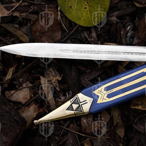 Pode incluir: Uma l&acirc;mina de espada prateada repousa sobre uma bainha azul com detalhes dourados. A bainha apresenta um design geom&eacute;trico dourado e uma ponta dourada. A espada e a bainha s&atilde;o colocadas sobre um fundo de folhas e galhos.