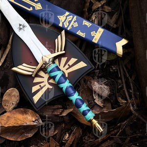 Pode incluir: R&eacute;plica detalhada de uma espada, escudo e bainha de fantasia. A espada apresenta uma l&acirc;mina prateada, detalhes dourados e uma empunhadura enrolada em azul e verde-azulado. O escudo &eacute; preto e vermelho com um emblema dourado.