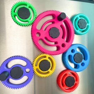 May include: A collection of colorful gear-shaped magnets in green, pink, blue, yellow, and red. Each gear has a black center and is attached to a metallic surface. The gears vary in size and are arranged in a visually appealing pattern.