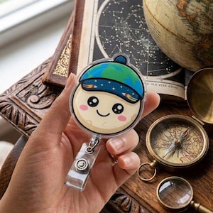 Puede incluir: Un portatarjetas redondo de estilo caricaturesco con un personaje de luna sonriente que lleva un sombrero con el tema de la Tierra azul y verde. El portatarjetas tiene un clip retráctil de plástico transparente. El fondo incluye un globo terráqueo vintage, una brújula y libros antiguos.