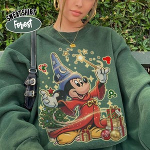 Peut inclure: Un sweat-shirt à col rond vert foncé avec une illustration d'une souris de dessin animé portant un chapeau de magicien bleu et une robe rouge, tenant une baguette dorée. Le motif inclut un sapin de Noël, des cadeaux et des étoiles dorées.