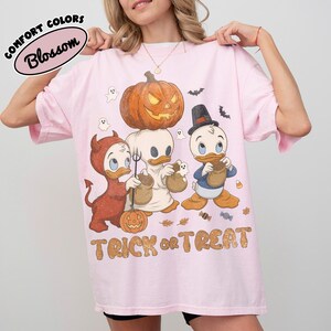 May include: A light pink t-shirt featuring a graphic of three cartoon ducks in Halloween costumes, including a devil, a ghost, and a top-hat wearer. A large orange jack-o'-lantern sits above them, with the text TRICK OR TREAT printed below.