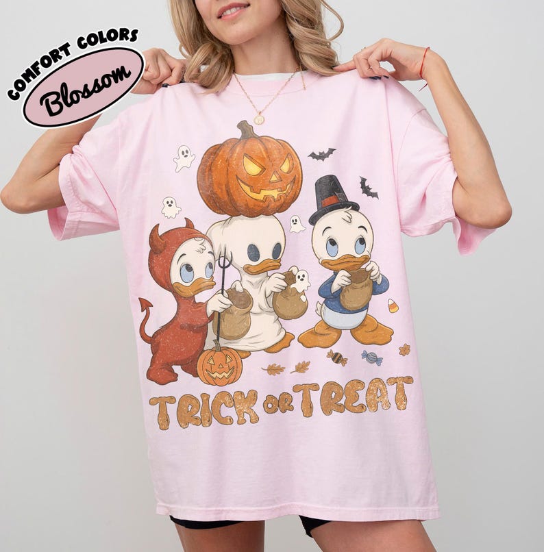 May include: A light pink t-shirt featuring a graphic of three cartoon ducks in Halloween costumes, including a devil, a ghost, and a top-hat wearer. A large orange jack-o'-lantern sits above them, with the text TRICK OR TREAT printed below.