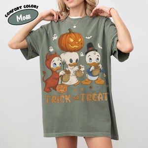 May include: A moss green t-shirt featuring a graphic of three cartoon ducks in Halloween costumes, including a devil, a ghost, and a top hat wearer, with a large jack-o'-lantern above them and the text TRICK or TREAT printed in orange letters.