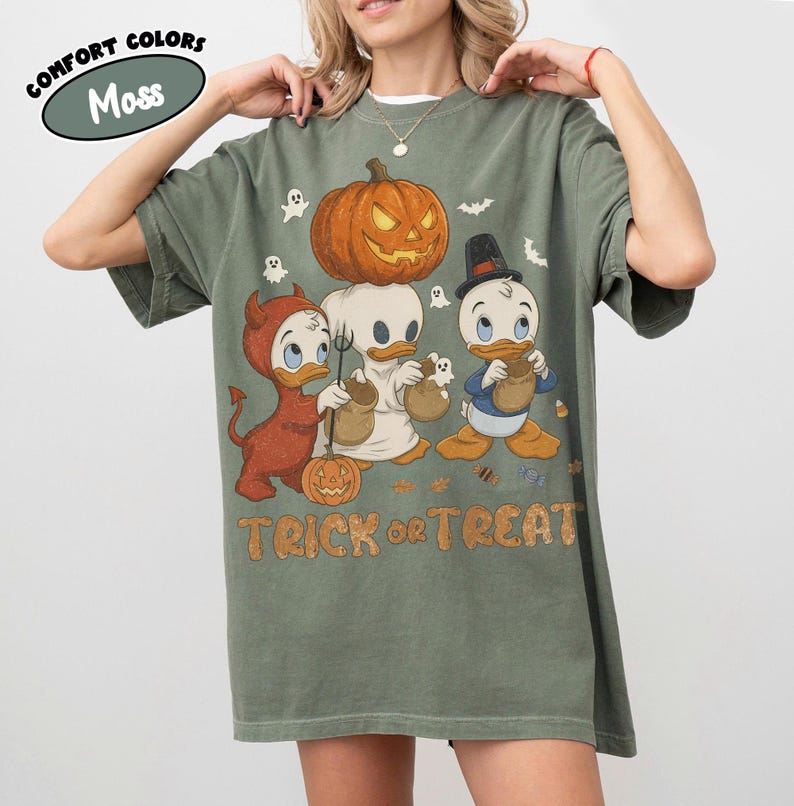 May include: A moss green t-shirt featuring a graphic of three cartoon ducks in Halloween costumes, including a devil, a ghost, and a top hat wearer, with a large jack-o'-lantern above them and the text TRICK or TREAT printed in orange letters.