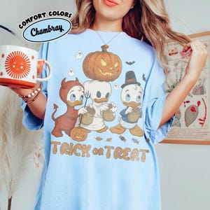 May include: A light blue t-shirt featuring a graphic of three cartoon ducks in Halloween costumes, including a devil, a ghost, and a pilgrim, with a large pumpkin, small ghosts, and the text TRICK or TREAT printed in a tan, textured font.