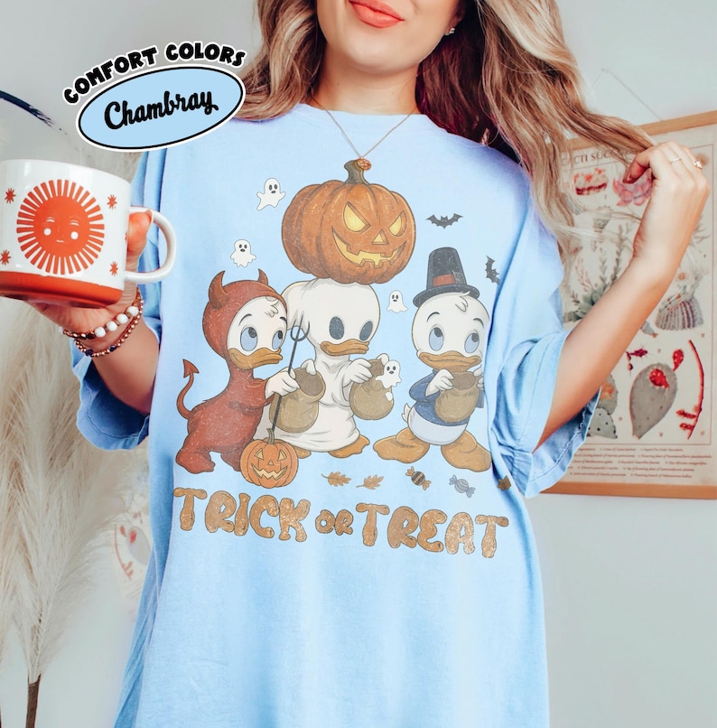 May include: A light blue t-shirt featuring a graphic of three cartoon ducks in Halloween costumes, including a devil, a ghost, and a pilgrim, with a large pumpkin, small ghosts, and the text TRICK or TREAT printed in a tan, textured font.