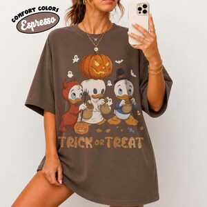 May include: An espresso brown short-sleeve t-shirt featuring a graphic of three cartoon ducks in Halloween costumes. The design includes a large orange jack-o'-lantern, small ghosts, and the text "TRICK or TREAT" in a stylized orange font.