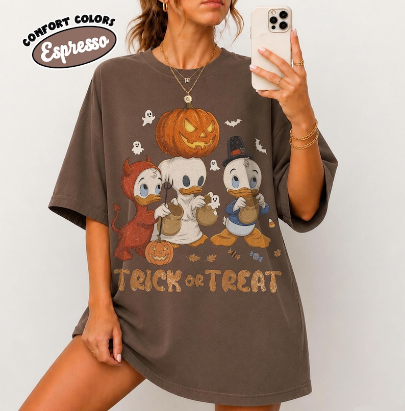 May include: An espresso brown short-sleeve t-shirt featuring a graphic of three cartoon ducks in Halloween costumes. The design includes a large orange jack-o'-lantern, small ghosts, and the text "TRICK or TREAT" in a stylized orange font.