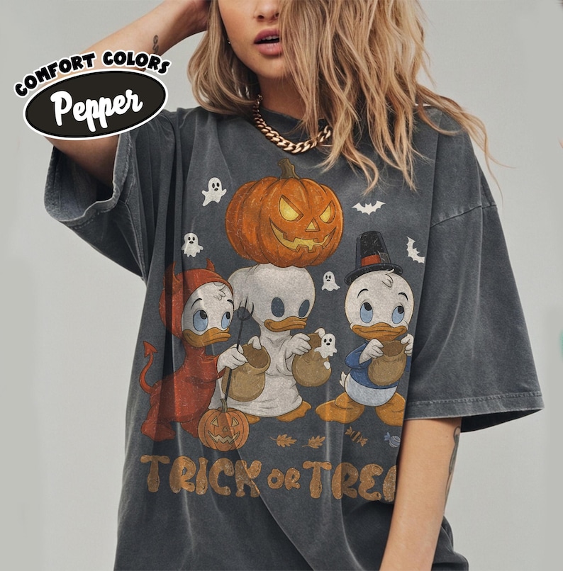 May include: A charcoal gray short-sleeve t-shirt featuring a graphic of three cartoon ducks in Halloween costumes, including a devil, a ghost, and a pilgrim, with a large pumpkin above them and the text TRICK OR TREAT printed at the bottom.
