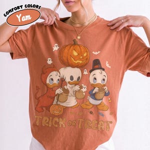 May include: A yam-colored t-shirt featuring a graphic of three cartoon ducks in Halloween costumes, including a devil, a ghost, and a pilgrim. A large jack-o'-lantern sits above them, and the text "TRICK or TREAT" is printed at the bottom.