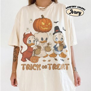 May include: An ivory t-shirt featuring a graphic of three cartoon ducks in Halloween costumes, including a devil, a ghost, and a pilgrim, with a large jack-o'-lantern above them. The text "TRICK or TREAT" is printed below in a brown, distressed font.