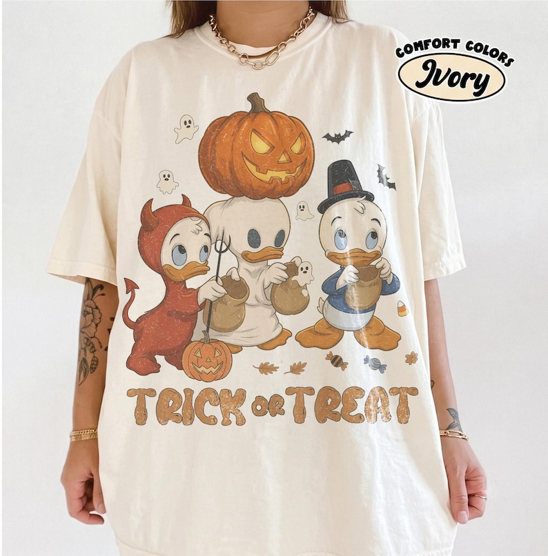 May include: An ivory t-shirt featuring a graphic of three cartoon ducks in Halloween costumes, including a devil, a ghost, and a pilgrim, with a large jack-o'-lantern above them. The text "TRICK or TREAT" is printed below in a brown, distressed font.