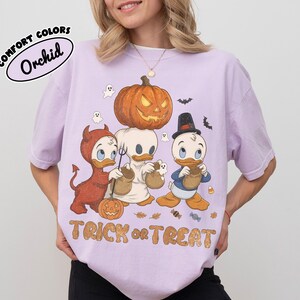 May include: A light purple t-shirt featuring a graphic of three cartoon ducks in Halloween costumes. One duck is dressed as a devil, one as a ghost, and one in a top hat. A large jack-o'-lantern sits above them, with the text TRICK or TREAT below.