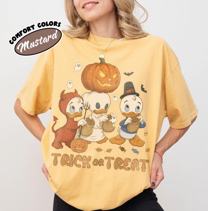 May include: A mustard yellow t-shirt featuring a graphic of three cartoon ducks in Halloween costumes, including a devil, a ghost, and a pilgrim. A large jack-o'-lantern sits atop the ghost's head. The text "TRICK or TREAT" is printed below.