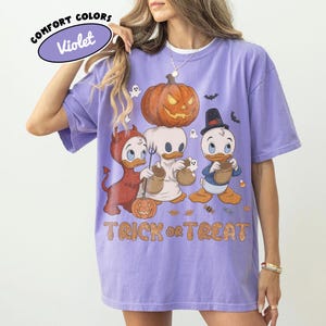 May include: A violet short-sleeve t-shirt featuring a graphic of three cartoon ducks in Halloween costumes, a large jack-o'-lantern, and the text "TRICK or TREAT". The design includes a devil, a ghost, and a wizard duck with candy bags.