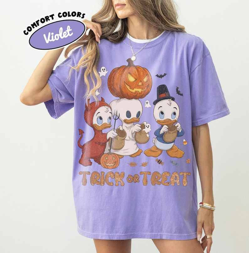May include: A violet short-sleeve t-shirt featuring a graphic of three cartoon ducks in Halloween costumes, a large jack-o'-lantern, and the text "TRICK or TREAT". The design includes a devil, a ghost, and a wizard duck with candy bags.