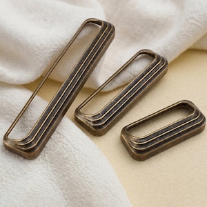 May include: Three antique bronze rectangular drawer pulls with a ridged design. The pulls are in varying sizes and are displayed on a white cloth and a light yellow surface. The hardware has a vintage aesthetic.