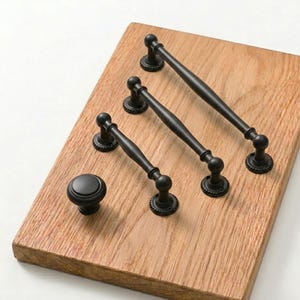Puede incluir: Un conjunto de herrajes negros para armarios, que incluye tres tiradores y un pomo, dispuestos sobre una superficie de madera marrón claro. Los tiradores tienen un diseño clásico con bases decorativas y extremos redondeados. El pomo es redondo y macizo.