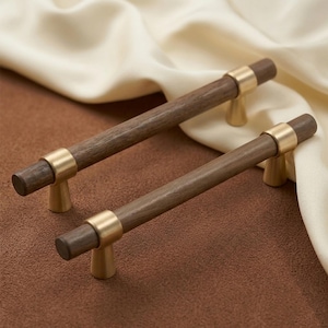 Puede incluir: Dos tiradores de armario de madera con acabado marrón oscuro y detalles en latón. Los tiradores tienen forma cilíndrica con un anillo de latón en el centro y una base de latón. Diseñados para muebles y armarios.