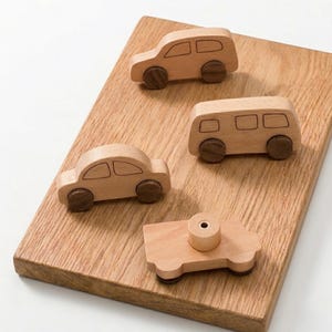 Puede incluir: Cuatro vehículos de juguete de madera: un coche, una furgoneta y otros dos modelos de coche. Los juguetes son de color marrón claro con ruedas marrones oscuras, colocados sobre una tabla de madera marrón claro. Los juguetes están diseñados con líneas simples y limpias.