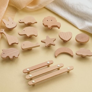 Puede incluir: Una colección de pomos y tiradores de madera para armarios en varias formas, incluyendo animales, estrellas y una luna. Los pomos son de color marrón claro y presentan una veta de madera natural. Los tiradores son rectangulares con detalles blancos.