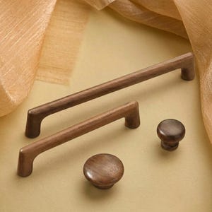 Può includere: Una collezione di ferramenta per mobili in legno, tra cui due maniglie e due pomelli. Le maniglie sono rettangolari con bordi arrotondati, e i pomelli sono circolari. Il legno ha una calda tonalità marrone medio. La ferramenta è esposta su una superficie beige chiaro.