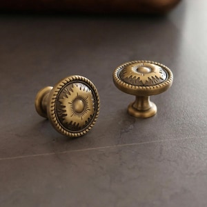 Puede incluir: Dos tiradores de armario de latón antiguo con un diseño decorativo de rayos de sol. Los tiradores tienen forma redonda con un borde similar a una cuerda. Uno está sobre un pedestal corto, el otro es plano.