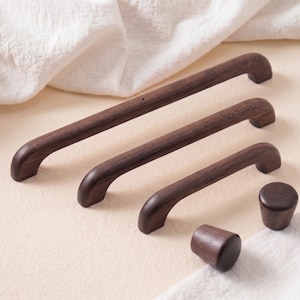 Può includere: Una collezione di ferramenta per mobili in legno marrone scuro, tra cui tre maniglie di diverse lunghezze e due pomelli. Le maniglie hanno un design curvo e i pomelli sono cilindrici. Gli articoli sono esposti su una superficie beige chiaro con tessuto bianco.
