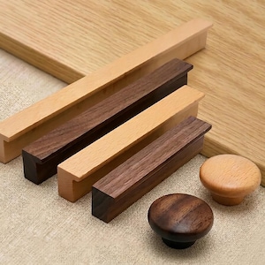 Puede incluir: Un conjunto de herrajes de madera para armarios, que incluye tiradores rectangulares y pomos redondos. Los tiradores son de tonos de madera claros y oscuros, y los pomos son de color marrón claro y oscuro. Los herrajes se muestran sobre una superficie beige claro.