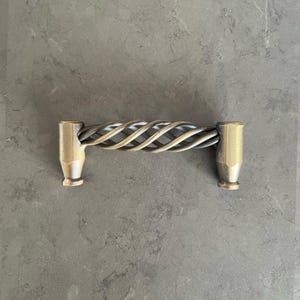 以下が含まれることがあります： ねじれた金属デザインのブロンズ色のキャビネットプル。ハンドルは、一連の織り交ぜられた金属ストランドを備えた円筒形をしています。プルの端は、フレアベースの無垢のブロンズ色です。キャビネットや引き出しに適しています。