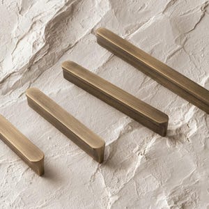 May include: Four antique brass cabinet pulls of varying lengths. The pulls are rectangular with rounded ends, and have a warm, golden-brown color. They are arranged diagonally on a textured, off-white surface.