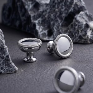 Könnte beinhalten: Ein Set silberner Manschettenknöpfe mit polierter, reflektierender Oberfläche und dekorativem, perlenbesetztem Rand. Die Manschettenknöpfe sind rund und liegen auf einer dunkelgrauen Oberfläche, mit einem kleinen Akzentstück im Sockelstil.