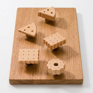 May include: Wooden cabinet knobs in the shape of cheese wedges, crackers, and a cookie, arranged on a wooden cutting board. The knobs are light brown with small, dark brown details.