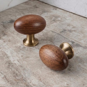 May include: Two wooden cabinet knobs with a brass base. The knobs have an oval shape and a rich brown color. One knob is upright, while the other is lying on its side. These are decorative hardware for furniture.