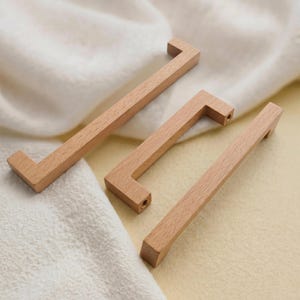 May include: Three wooden cabinet pulls with a natural wood grain finish. Each pull has a rectangular bar with an L-shaped handle. The pulls are arranged on a soft, off-white fabric.