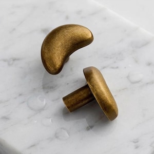 May include: Two brass cabinet knobs, each with a crescent shape and a cylindrical post. The knobs are a warm gold colour and rest on a white marble surface. These knobs are likely for a drawer or cabinet.