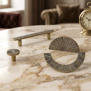 May include: A collection of antique brass cabinet hardware, including a long bar pull, a round knob, and two half-circle pulls. The hardware features a textured, linear design and is displayed on a marble surface. A vintage clock is in the background.