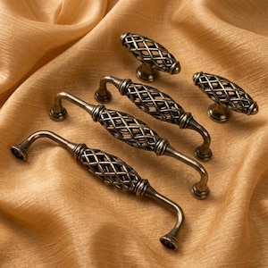 Puede incluir: Un surtido de herrajes de gabinete de bronce antiguo, que incluyen manijas y perillas. Las manijas tienen un diseño curvo con una jaula con patrón de diamantes, y las perillas tienen un diseño de jaula similar. El hardware se muestra sobre una tela dorada.