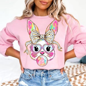 May include: Pink sweatshirt featuring a cartoon bunny face with heart-shaped glasses and a bubblegum bubble. The bunny has a bow with black polka dots and colorful ears. The sweatshirt is a solid light pink color.