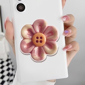 May include: A white phone case featuring a large, three-dimensional flower design. The flower has pink and cream-colored petals with a textured appearance, and an orange button center with four small holes.
