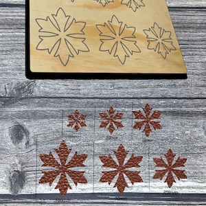 Op de afbeelding: Een houten stanssjabloon met sneeuwvlokontwerpen. De sjabloon heeft sneeuwvlokvormen in verschillende maten, de grootste meet 10 cm. De uitgestanste sneeuwvlokken zijn bruin en hebben een textuur. De sjabloon ligt op een houten oppervlak.