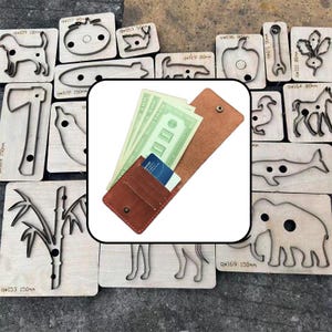 May include: A brown leather wallet with a snap closure, containing several green paper bills and a blue credit card, positioned over a background of various wooden cutting dies featuring animal and object shapes with metal outlines.