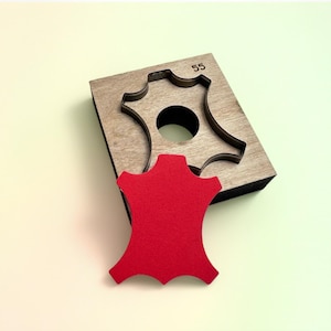Op de afbeelding: Een houten stansgereedschap met een lederen vorm en het nummer 55 gegraveerd op de bovenkant. Een rood leren stuk ligt naast het stansgereedschap. De tool wordt gebruikt voor het snijden van leer.