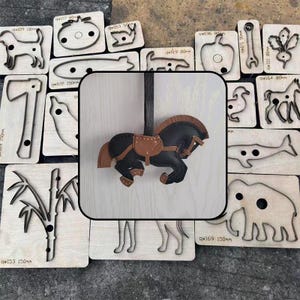 Op de afbeelding: Een zwart en bruin leren paardenornament met een zadel, omringd door houten stansvormen van diverse objecten. Het paard is ongeveer 10 cm lang. Andere vormen zijn een bijl, een hond en een olifant.