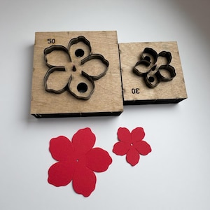 Op de afbeelding: Twee houten bloemvormige stansblokken met metalen contouren, gemarkeerd met "50" en "30", liggen boven twee rode viltbloemuitsparingen. De stansvormen zijn voor het knutselen en creëren van bloemmotieven.