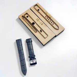 以下が含まれることがあります： 「16-14」の文字が入った木製の時計ストラップカッティングテンプレートと、クロコダイル柄の濃い青色の革製時計ストラップ2本。1つのストラップにはシルバーのバックルが付いています。テンプレートは明るい色の木材で作られています。